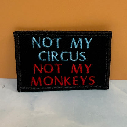 A rectangular black embroidered patch with the text 'NOT MY CIRCUS NOT MY MONKEYS' in light blue and red thread, displayed on a white marble-like surface against an orange background.