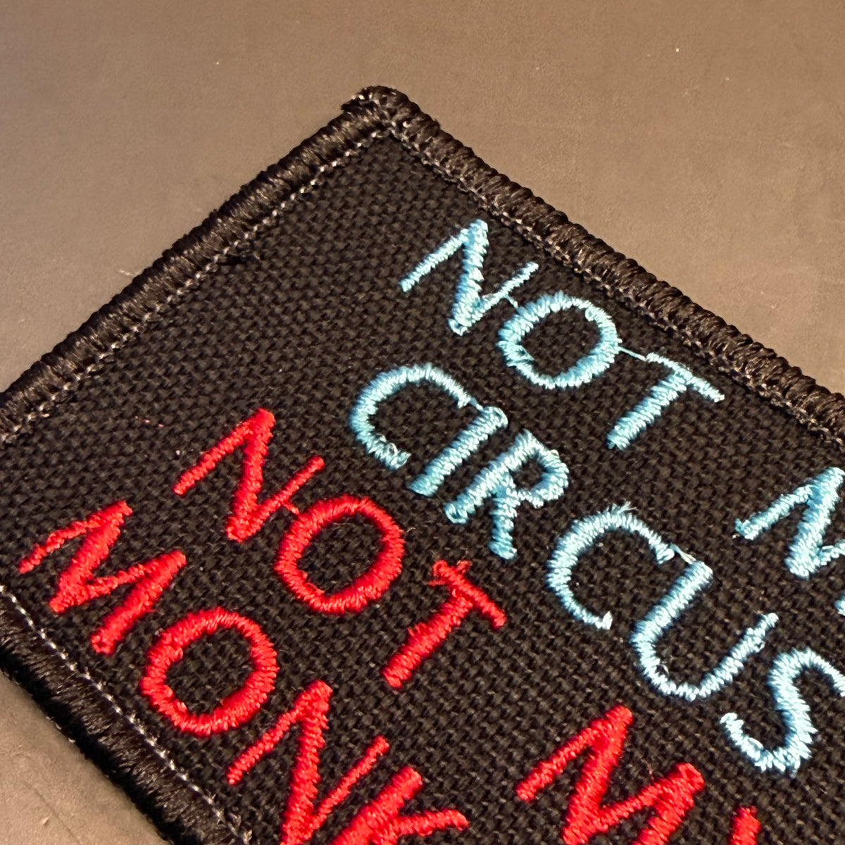 A close-up of a black embroidered patch with the phrase 'NOT A CIRCUS MONKEY' in red, light blue, and white embroidery on a dark brown background.