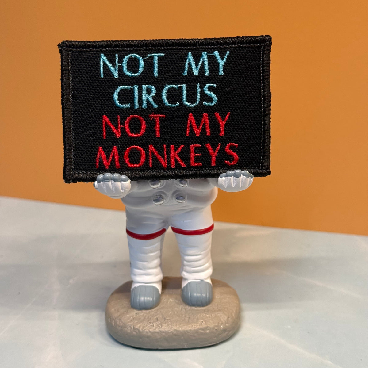 A small figurine of an astronaut holding a black sign that reads 'NOT MY CIRCUS NOT MY MONKEYS' in light blue and red embroidery, standing on a round base against an orange background.