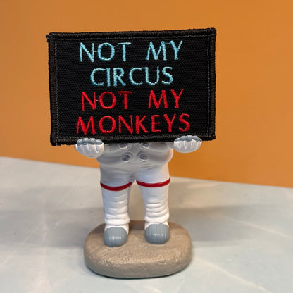 A small figurine of an astronaut holding a black sign that reads 'NOT MY CIRCUS NOT MY MONKEYS' in light blue and red embroidery, standing on a round base against an orange background.