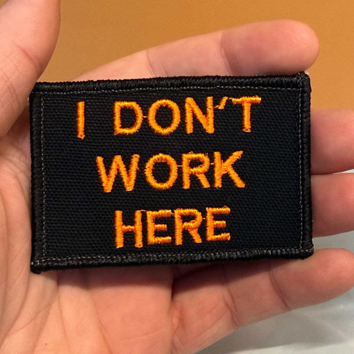 A rectangular black embroidered patch with orange text reading 'I DON'T WORK HERE', held in a person's hand against a warm tan background.