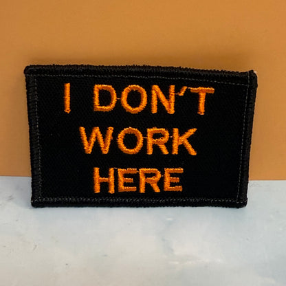 A rectangular black embroidered patch with the text 'I DON'T WORK HERE' in bright orange lettering, displayed on a white marble-like surface against a solid orange background.