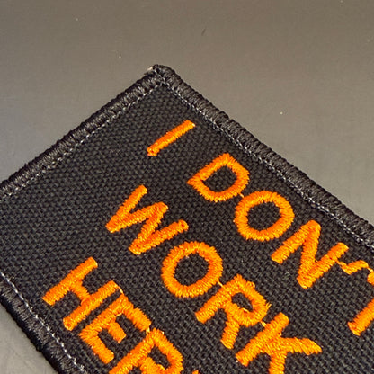 A close-up of a black embroidered patch with orange text reading 'I DON'T WORK HERE' on a dark gray background.