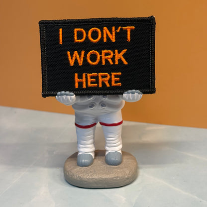A novelty astronaut figurine holding a black rectangular sign with orange embroidered text that reads 'I DON'T WORK HERE'. The figurine has a white spacesuit with red stripes and gray boots, standing on a gray rock base against an orange background.