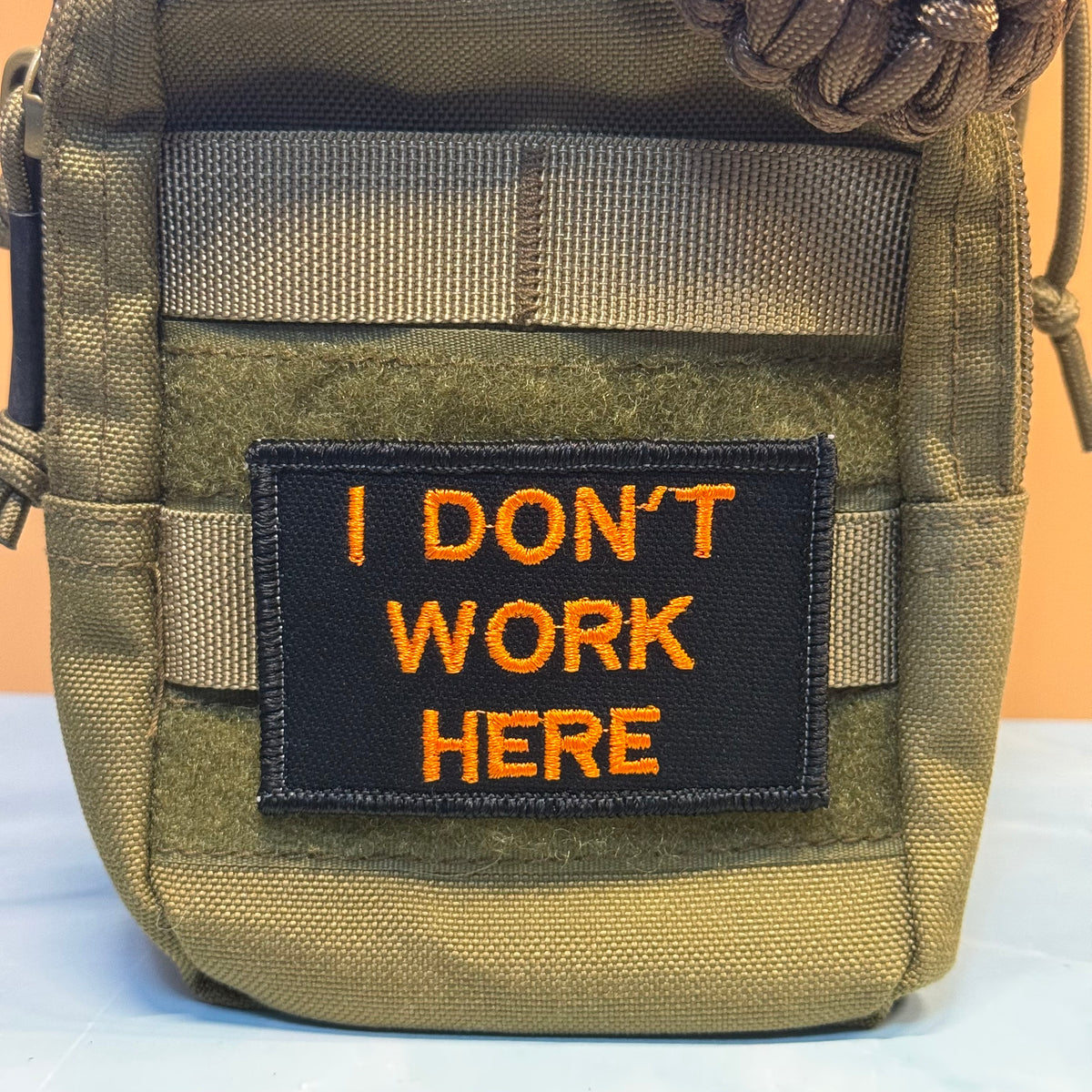 A close-up of a tactical pouch in olive drab and coyote brown webbing, featuring a black rectangular patch with orange embroidered text that reads 'I DON'T WORK HERE'.