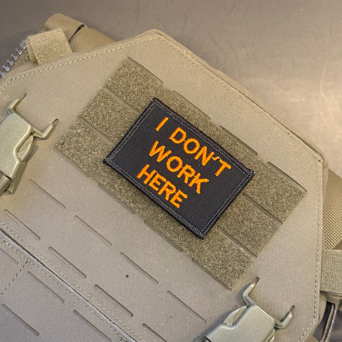 A rectangular black tactical patch with orange embroidered text reading 'I DON'T WORK HERE', attached to tan webbing on a beige tactical bag or vest.