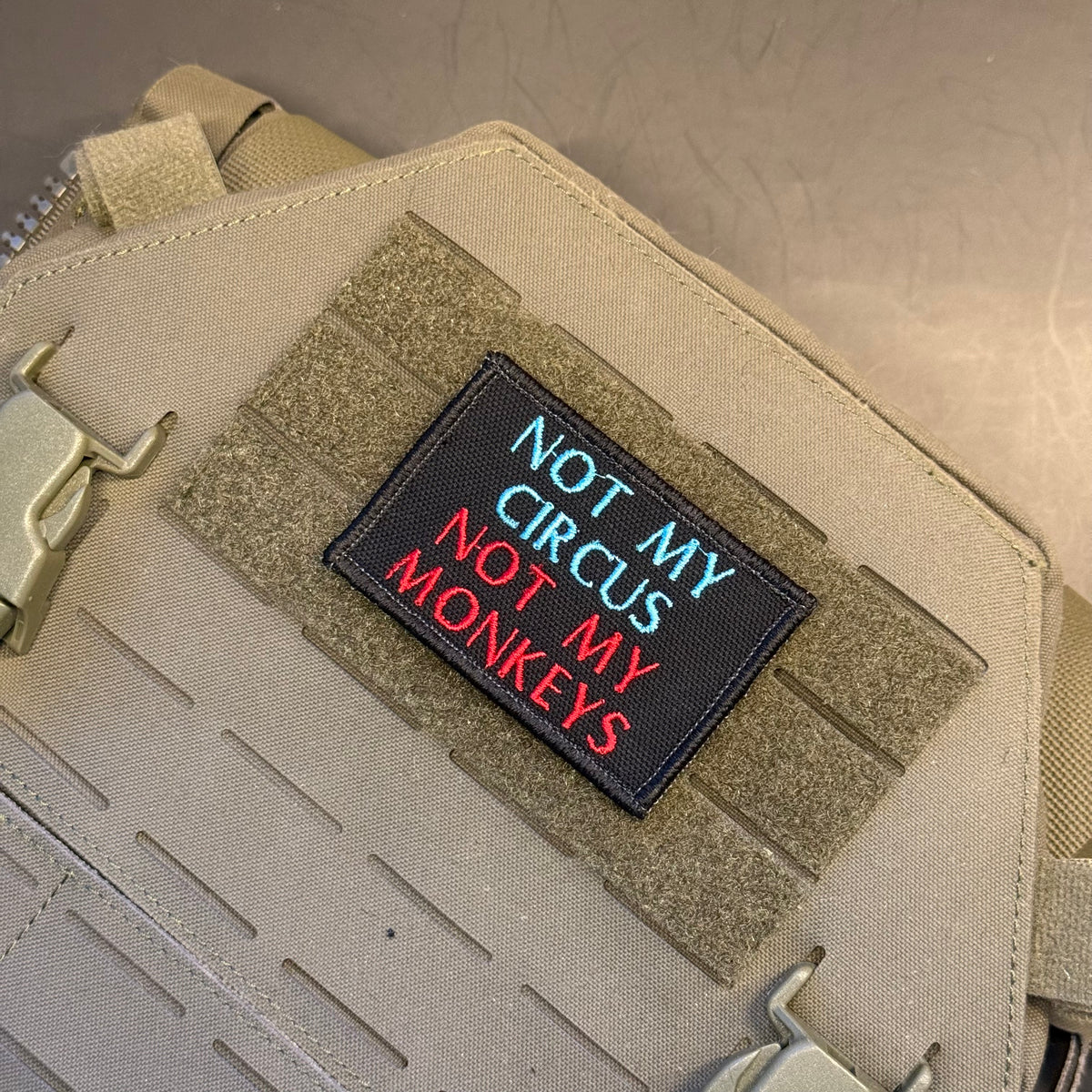 A rectangular black embroidered patch with the text 'NOT MY CIRCUS NOT MY MONKEYS' in blue and red lettering, shown attached to tan tactical webbing on a beige bag or gear panel.