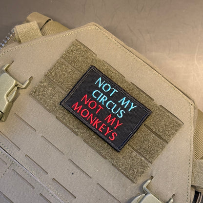 A rectangular black embroidered patch with the text 'NOT MY CIRCUS NOT MY MONKEYS' in blue and red lettering, shown attached to tan tactical webbing on a beige bag or gear panel.