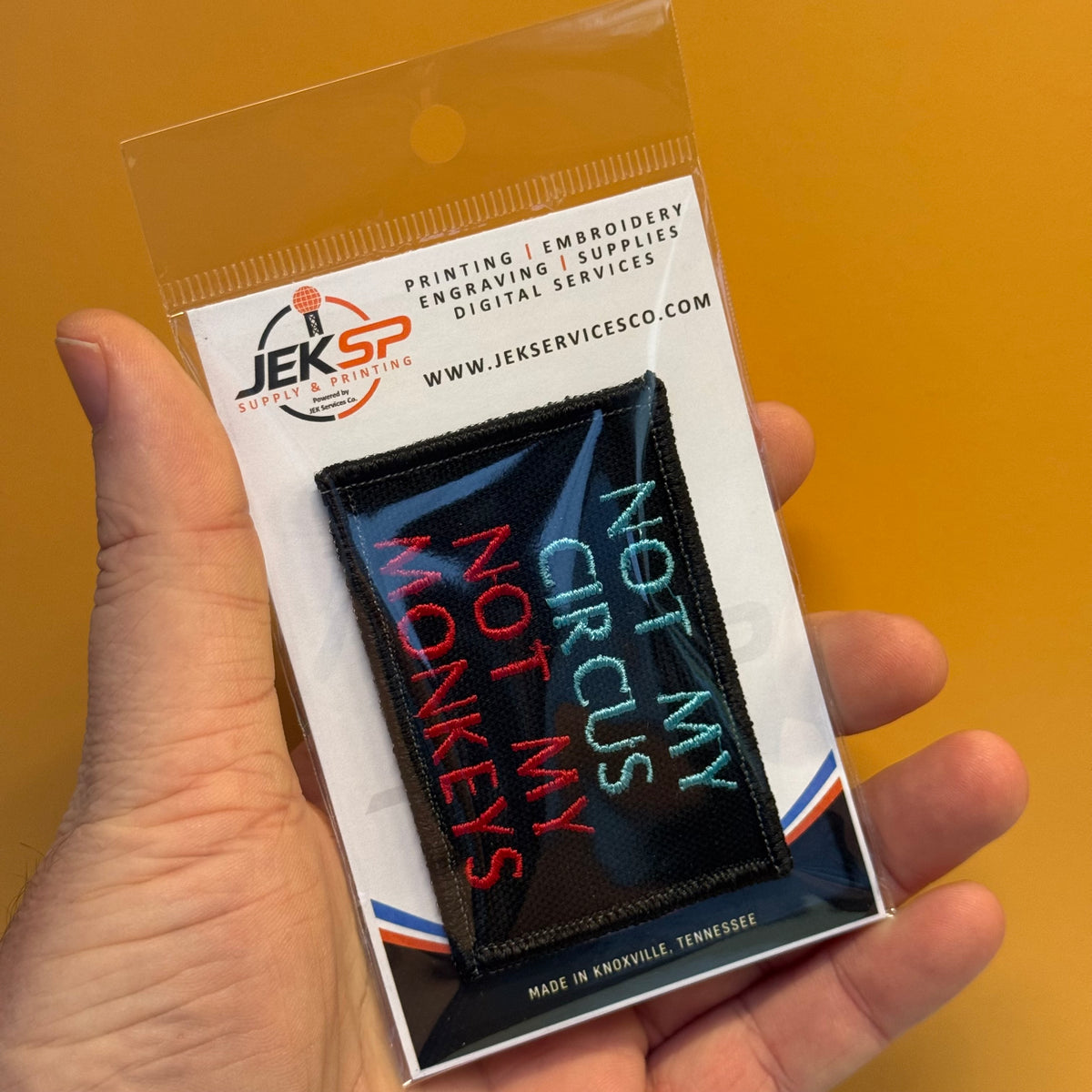A hand holds a packaged embroidered patch that reads 'NOT MY CIRCUS NOT MY MONKEYS' in red, teal, and white on a black background. The packaging shows the brand JEKSP Embroidery Supply.
