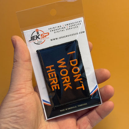 A hand holds a clear plastic blister pack containing a black embroidered patch with orange text reading 'I DON'T WORK HERE.' The package card shows the JEKSP branding and website, against a solid warm orange background.