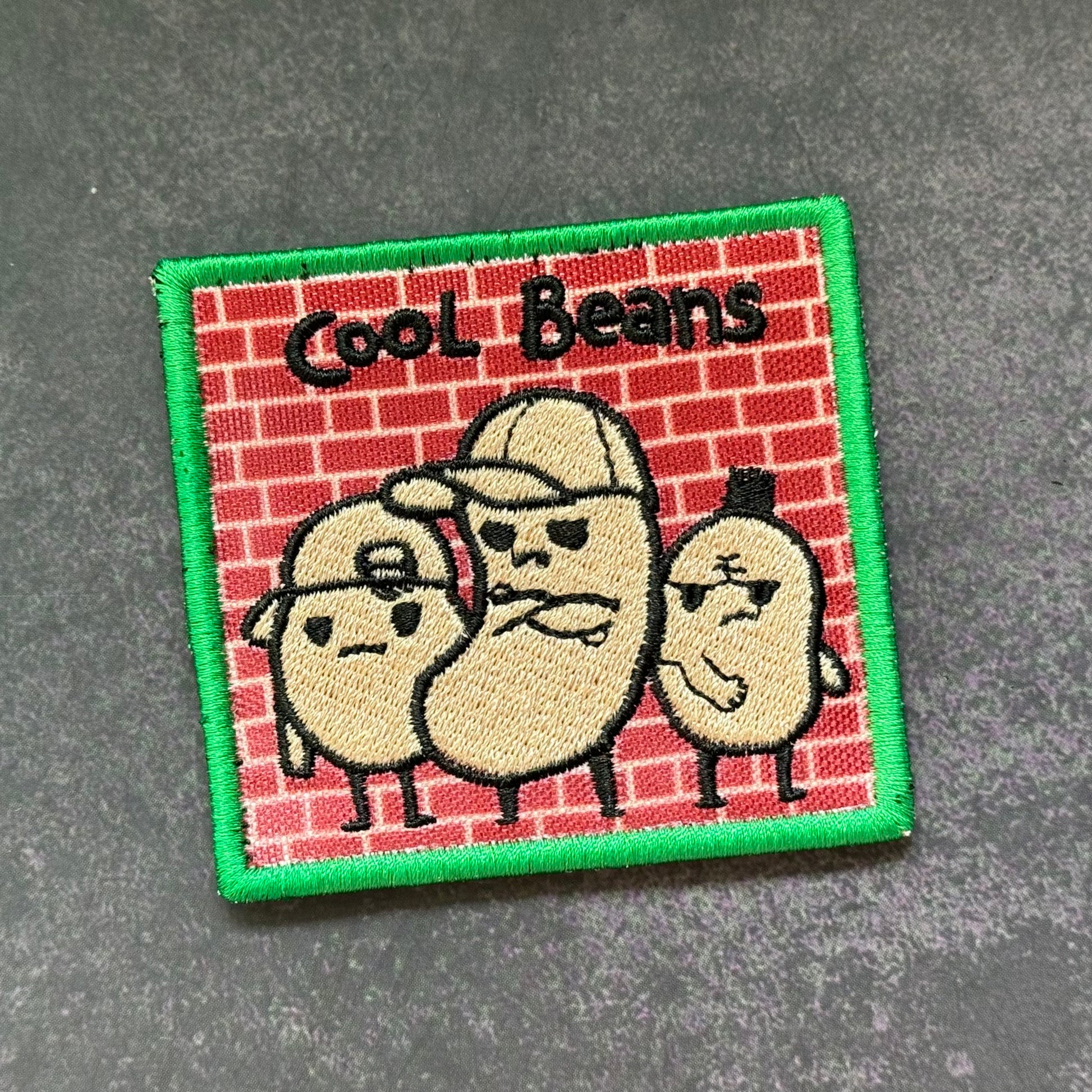 A square embroidered patch featuring three cartoon beans wearing caps against a red brick wall background, with the text 'Cool Beans' at the top and a green stitched border, shown on a dark gray textured surface.