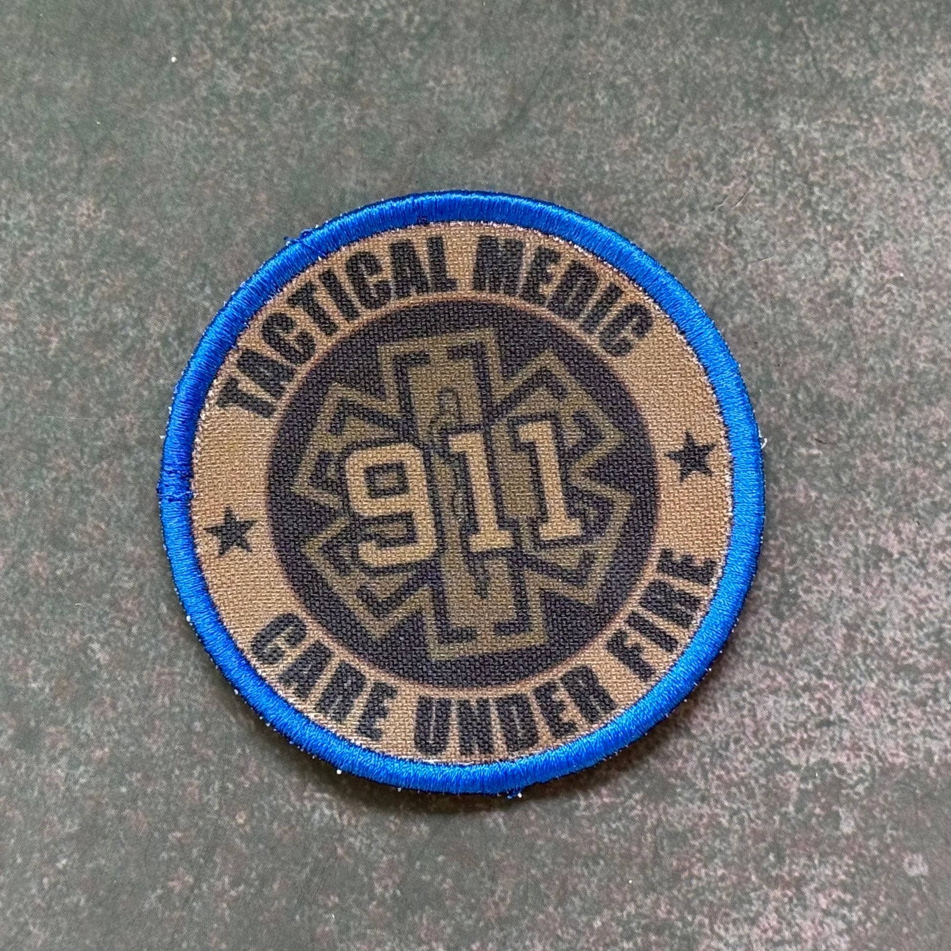 A circular tactical medic patch with a blue border, tan background, and a dark center featuring the text 'TACTICAL MEDIC', '911', and 'CARE UNDER FIRE' on a textured gray surface.