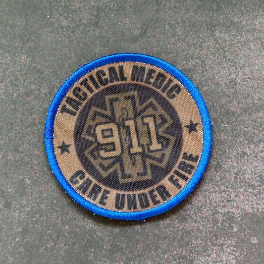 A circular tactical medic patch with a blue border, tan background, and a dark center featuring the text 'TACTICAL MEDIC', '911', and 'CARE UNDER FIRE' on a textured gray surface.