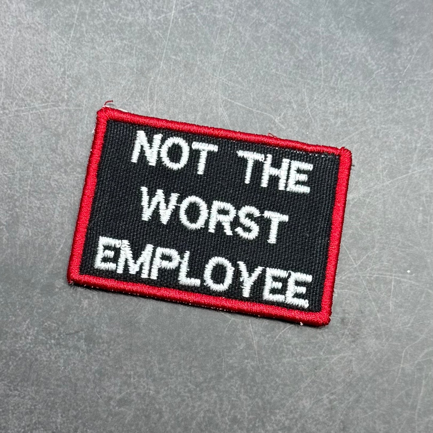 Black embroidered morale patch with a red border and white text reading "Not The Worst Employee" on a gray background.