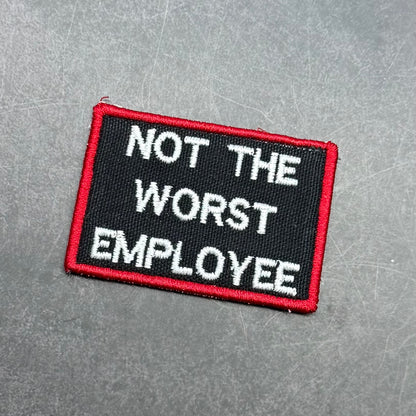 Black embroidered morale patch with a red border and white text reading "Not The Worst Employee" on a gray background.