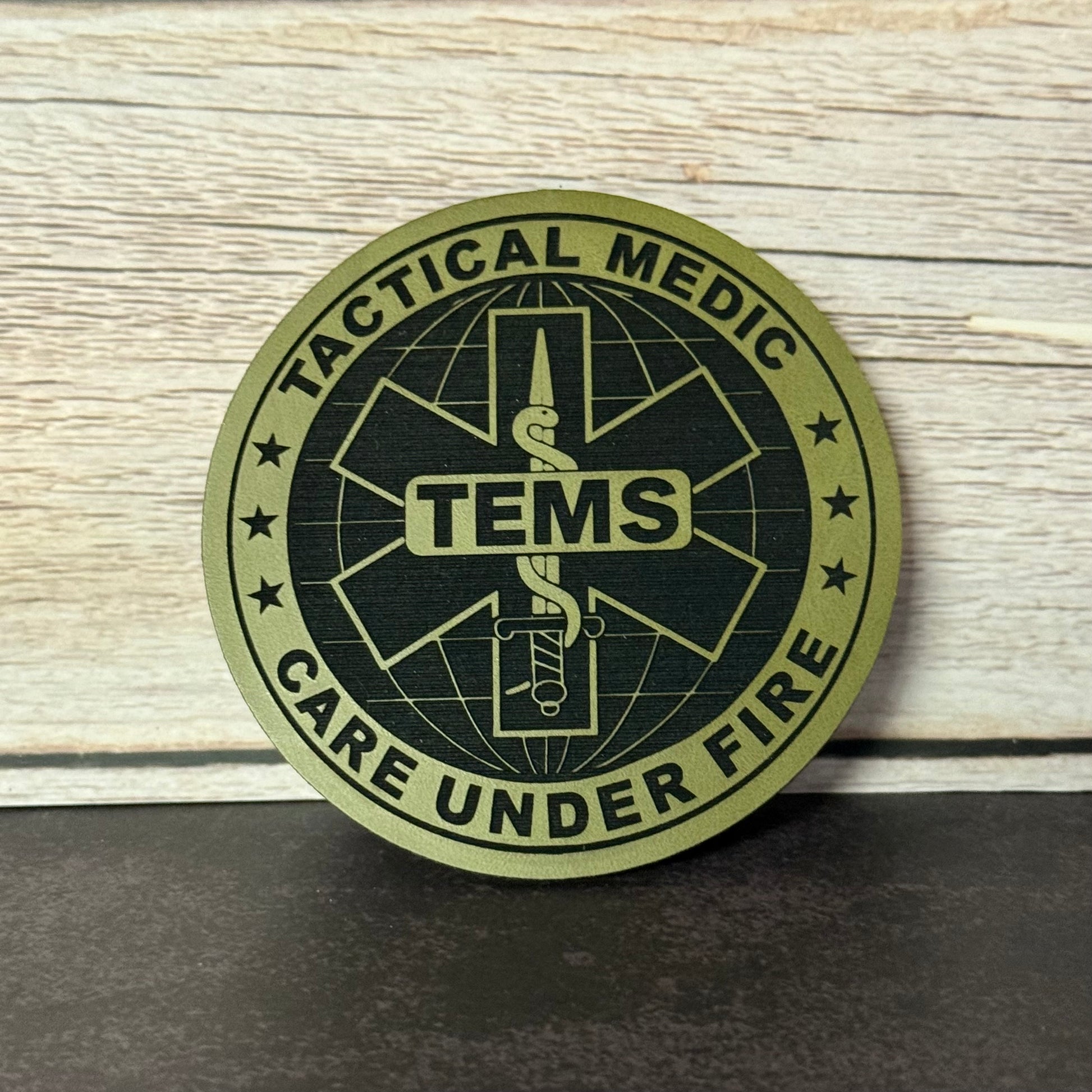 A circular tactical medic patch with a black and olive green design featuring the text 'TACTICAL MEDIC' and 'CARE UNDER FIRE' around a central medical symbol with 'TEMS' in the center, displayed on a dark surface against a light wood background.