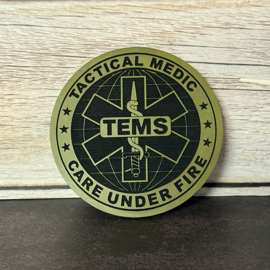A circular tactical medic patch with a black and olive green design featuring the text 'TACTICAL MEDIC' and 'CARE UNDER FIRE' around a central medical symbol with 'TEMS' in the center, displayed on a dark surface against a light wood background.