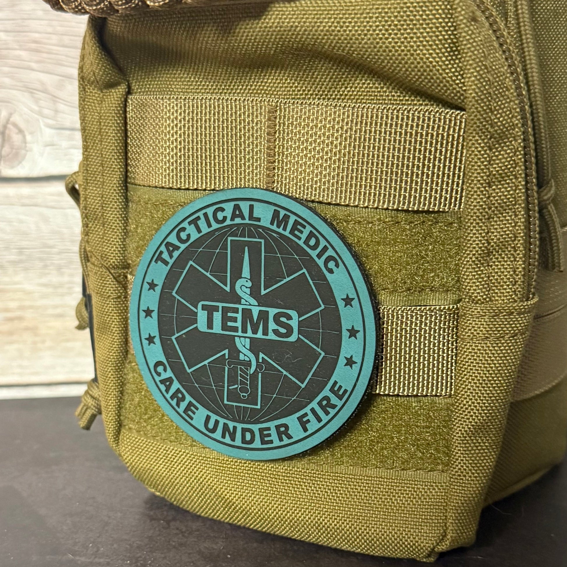 An olive drab tactical pouch with a rolled-up top showing a tan paracord cord, featuring a circular teal and black TENS 'Tactical Medic Care Under Fire' patch on the front, set against a light wood panel background.