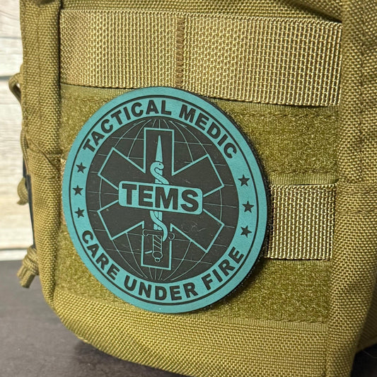 An olive drab tactical pouch with a rolled-up top showing a tan paracord cord, featuring a circular teal and black TENS 'Tactical Medic Care Under Fire' patch on the front, set against a light wood panel background.