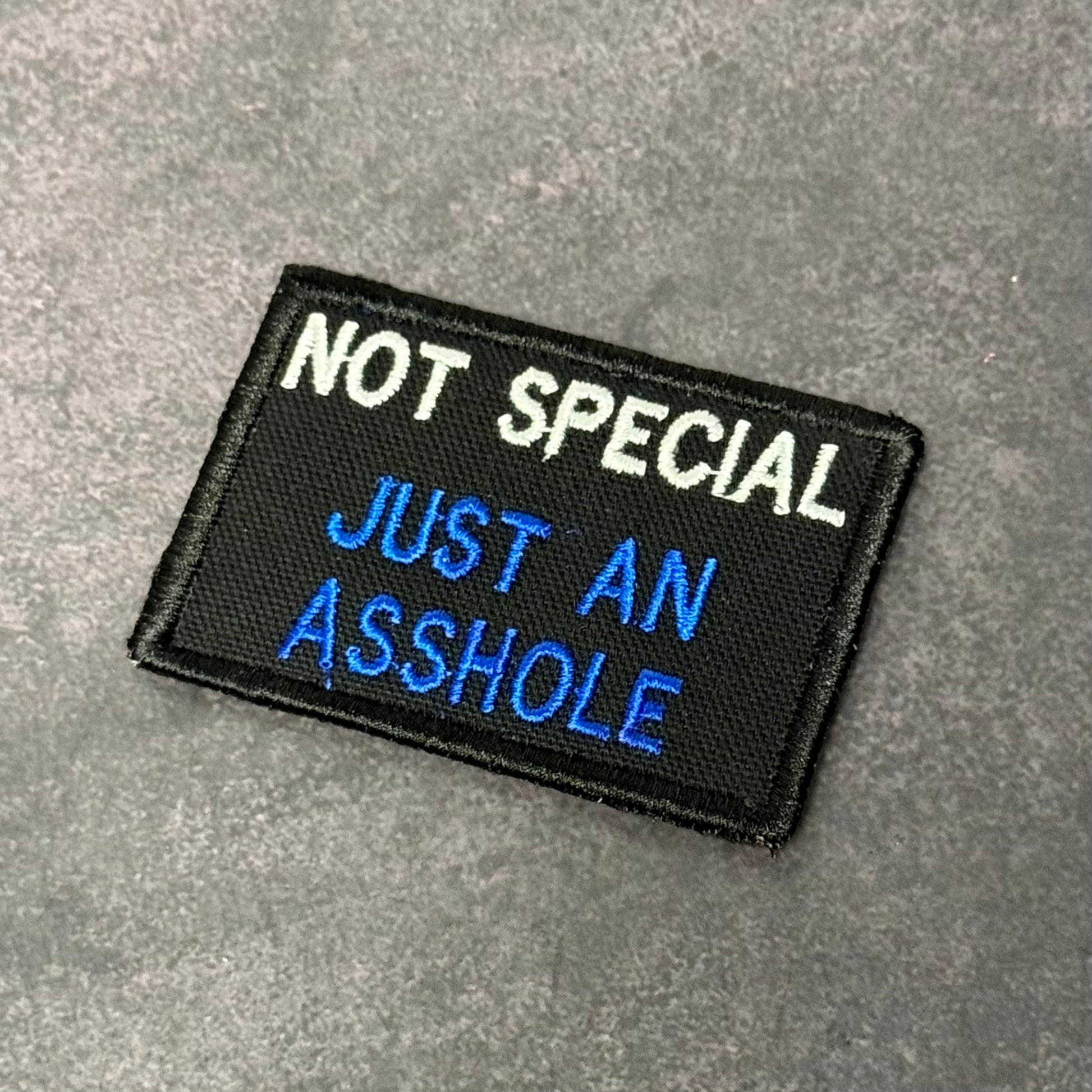 Black embroidered morale patch with white and blue text reading "Not Special Just An Asshole" displayed on a gray background.