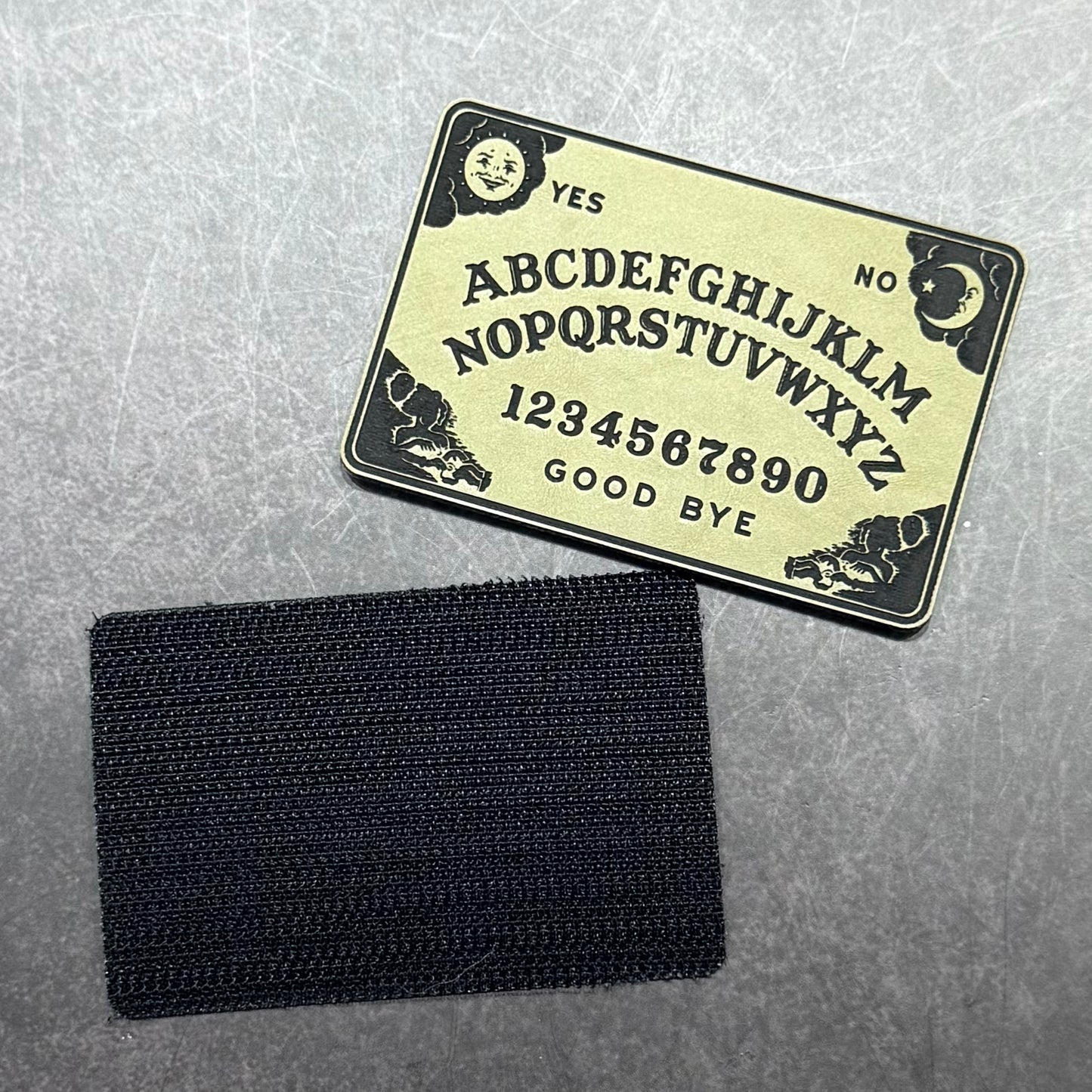 A black rectangular hook-and-loop patch shown below a black printed Ouija board template on a textured gray background.