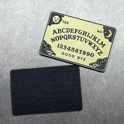 A black rectangular hook-and-loop patch shown below a black printed Ouija board template on a textured gray background.