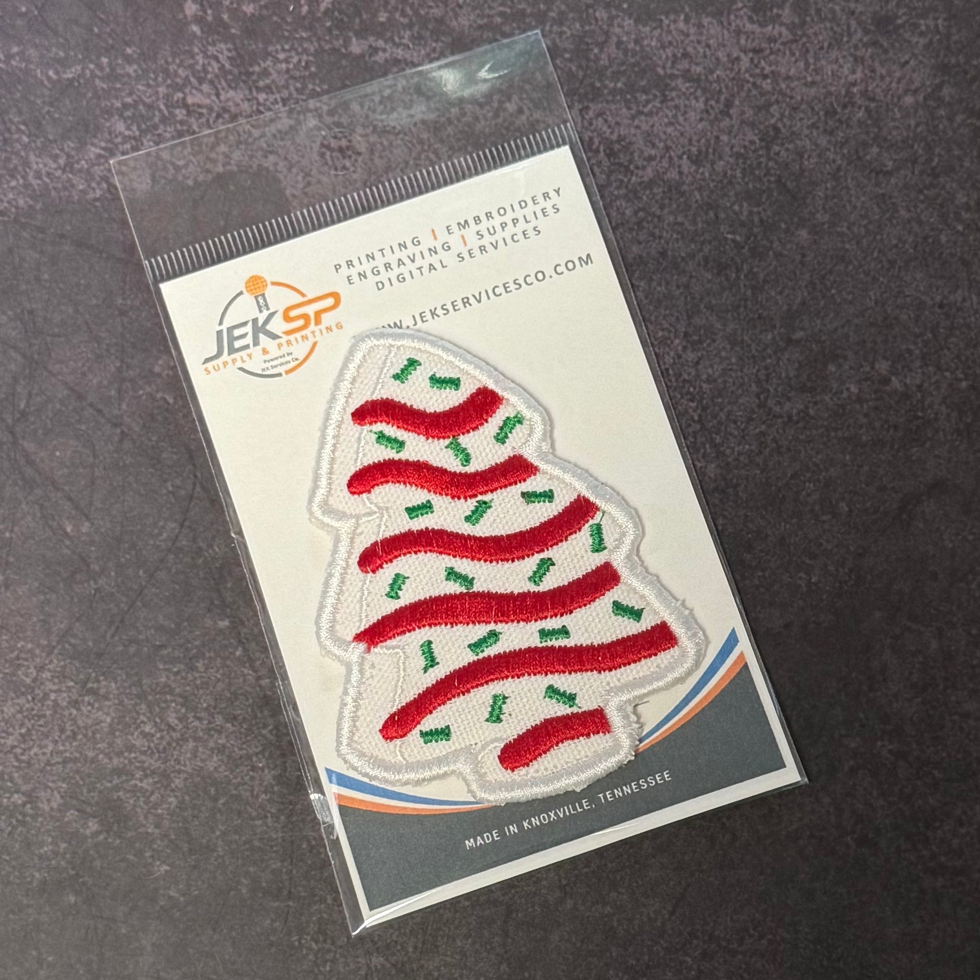 An embroidered Christmas tree patch in red and green on a white fabric base, sealed in a clear plastic sleeve with a printed backing card. The card shows the JEKSP embroidery services logo and text, and the patch is photographed on a dark textured surface.