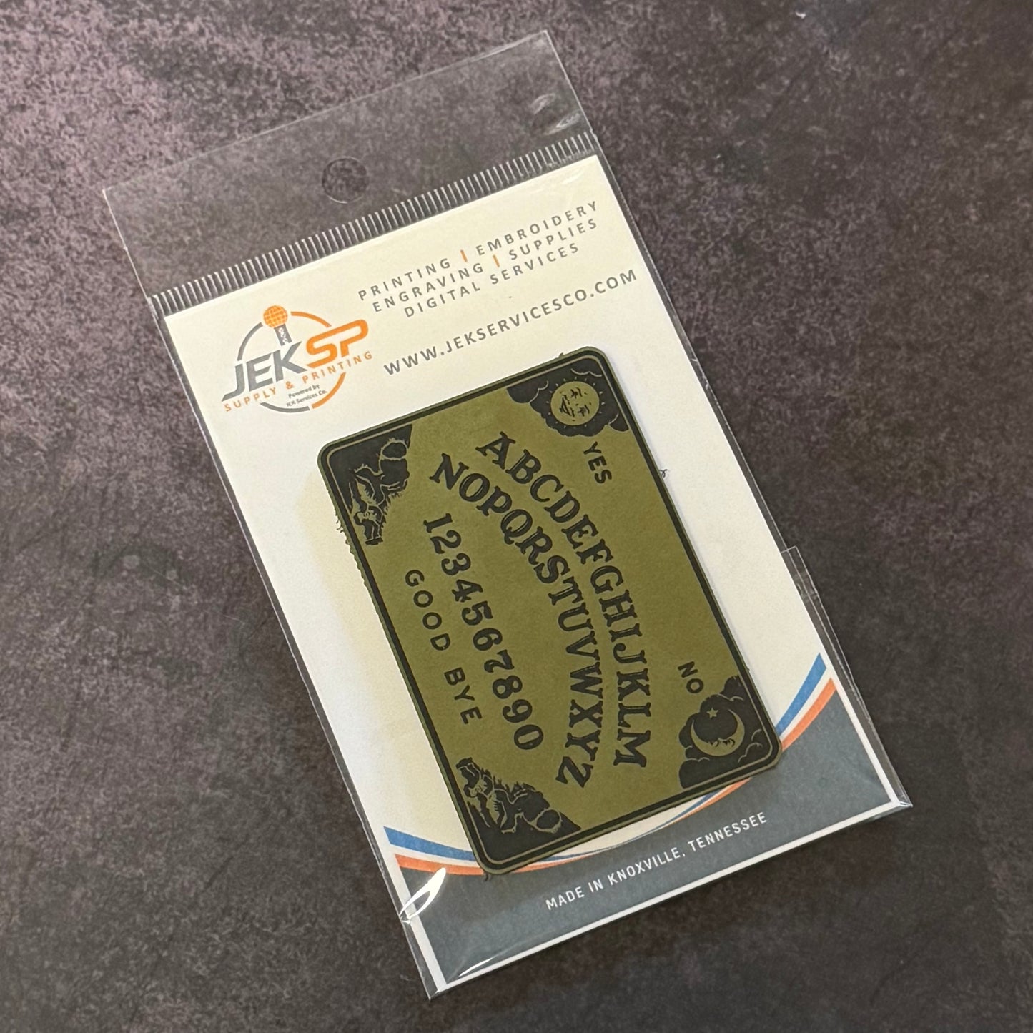 A printed Ouija board card inside a clear plastic sleeve, mounted on a branded backing card. The board is olive green with black lettering and symbols, photographed on a dark textured surface.