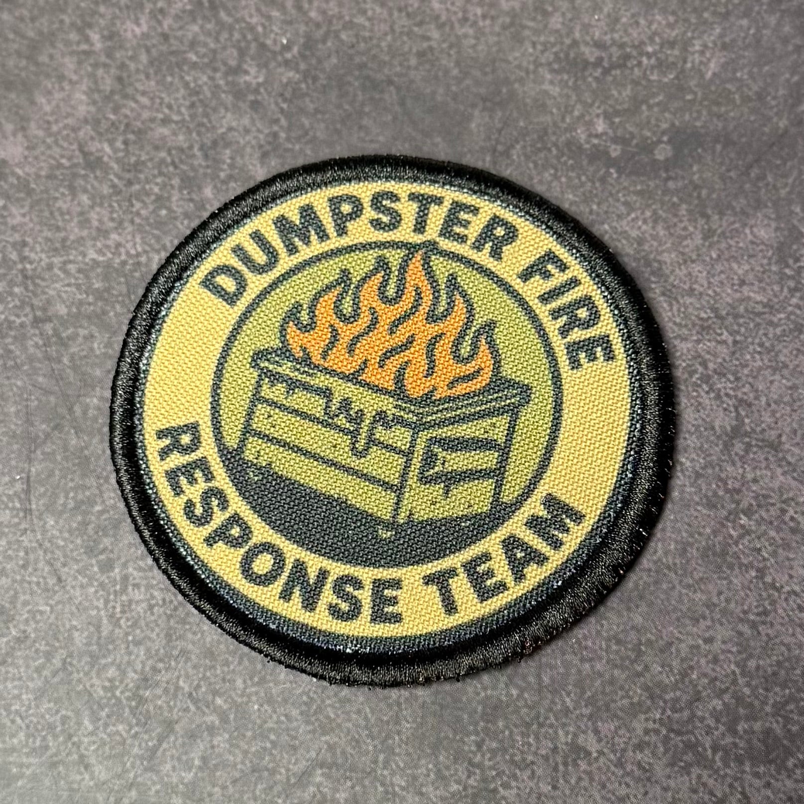 Round embroidered morale patch featuring a flaming dumpster graphic and the words "Dumpster Fire Response Team" on a tan and black background displayed on a gray textured surface.