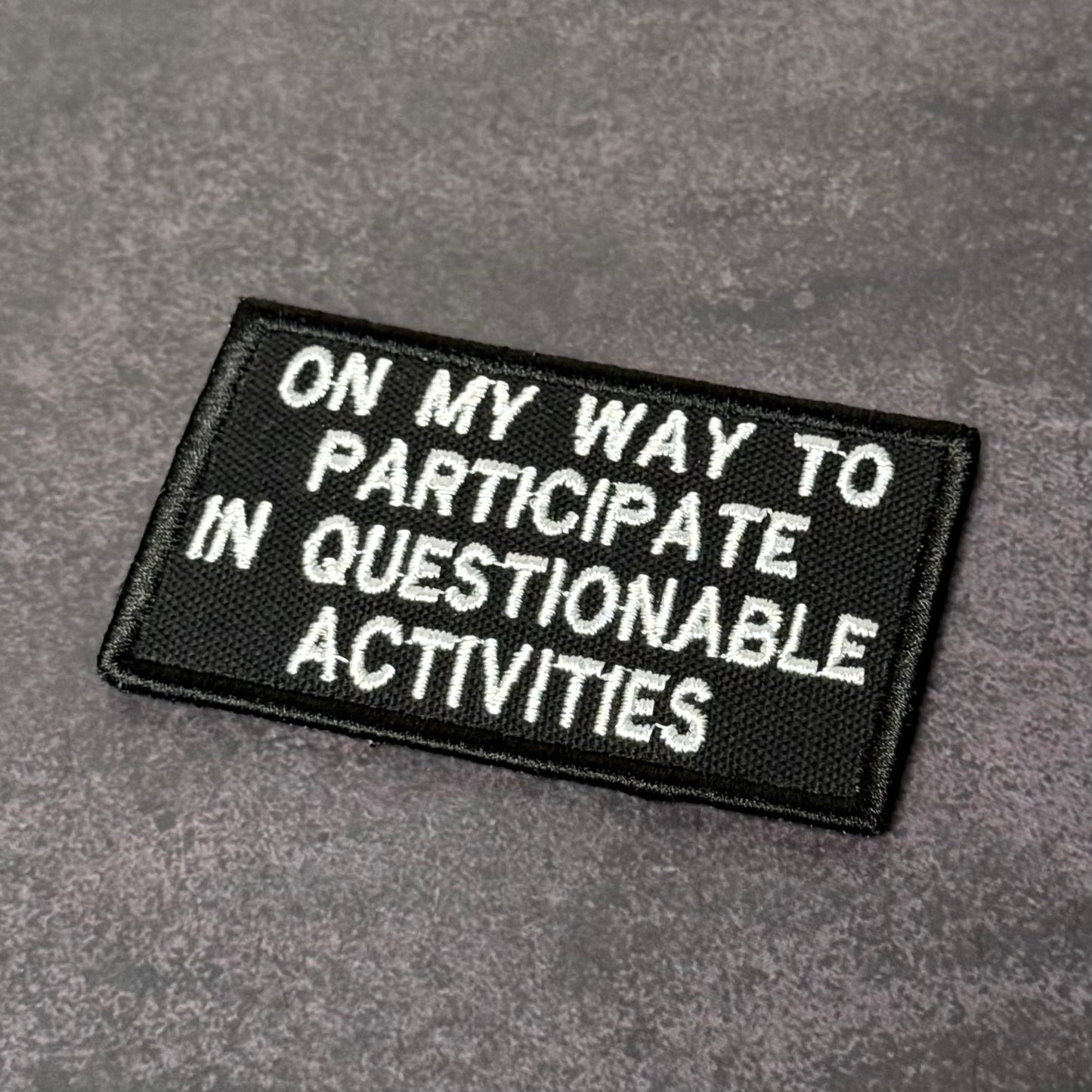 Black embroidered morale patch with white text reading "On My Way to Participate in Questionable Activities" displayed on a gray background.