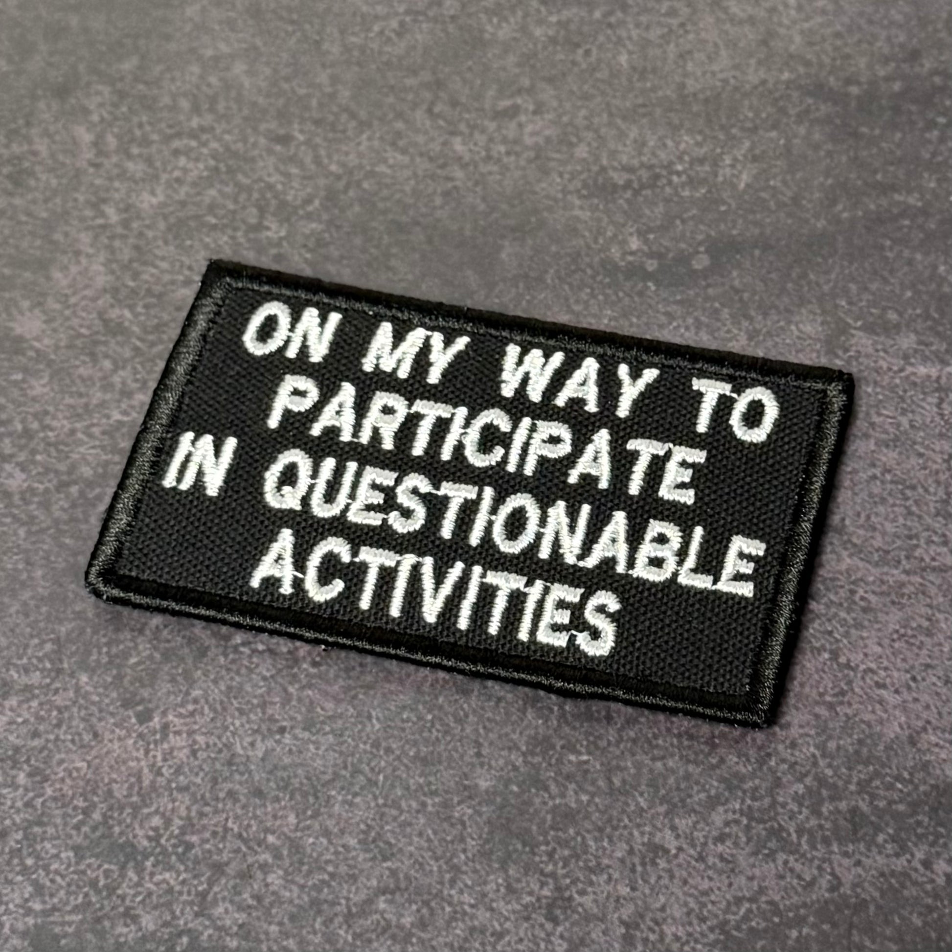 Black embroidered morale patch with white text reading "On My Way to Participate in Questionable Activities" displayed on a gray background.