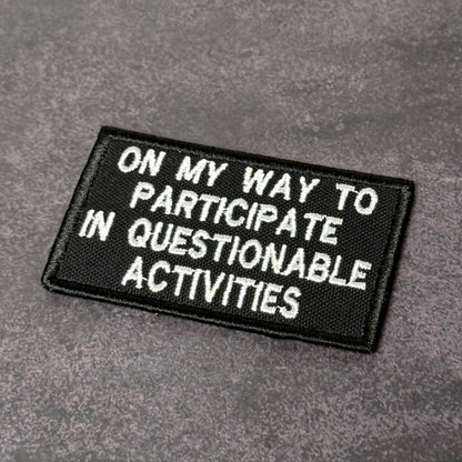 Black embroidered morale patch with white text reading "On My Way to Participate in Questionable Activities" displayed on a gray background.