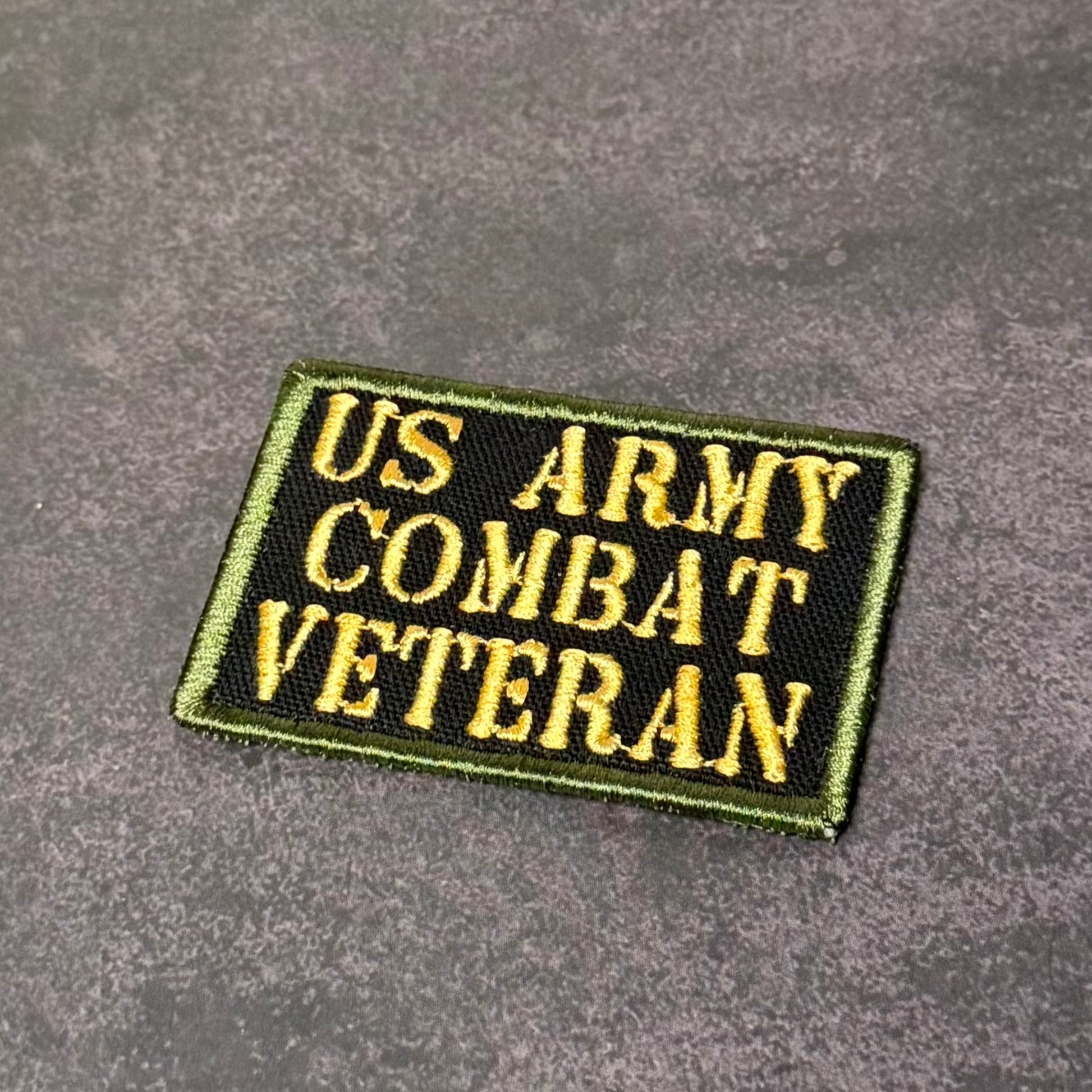 Black embroidered morale patch with gold lettering reading "US Army Combat Veteran" and an olive drab border displayed on a gray textured background.