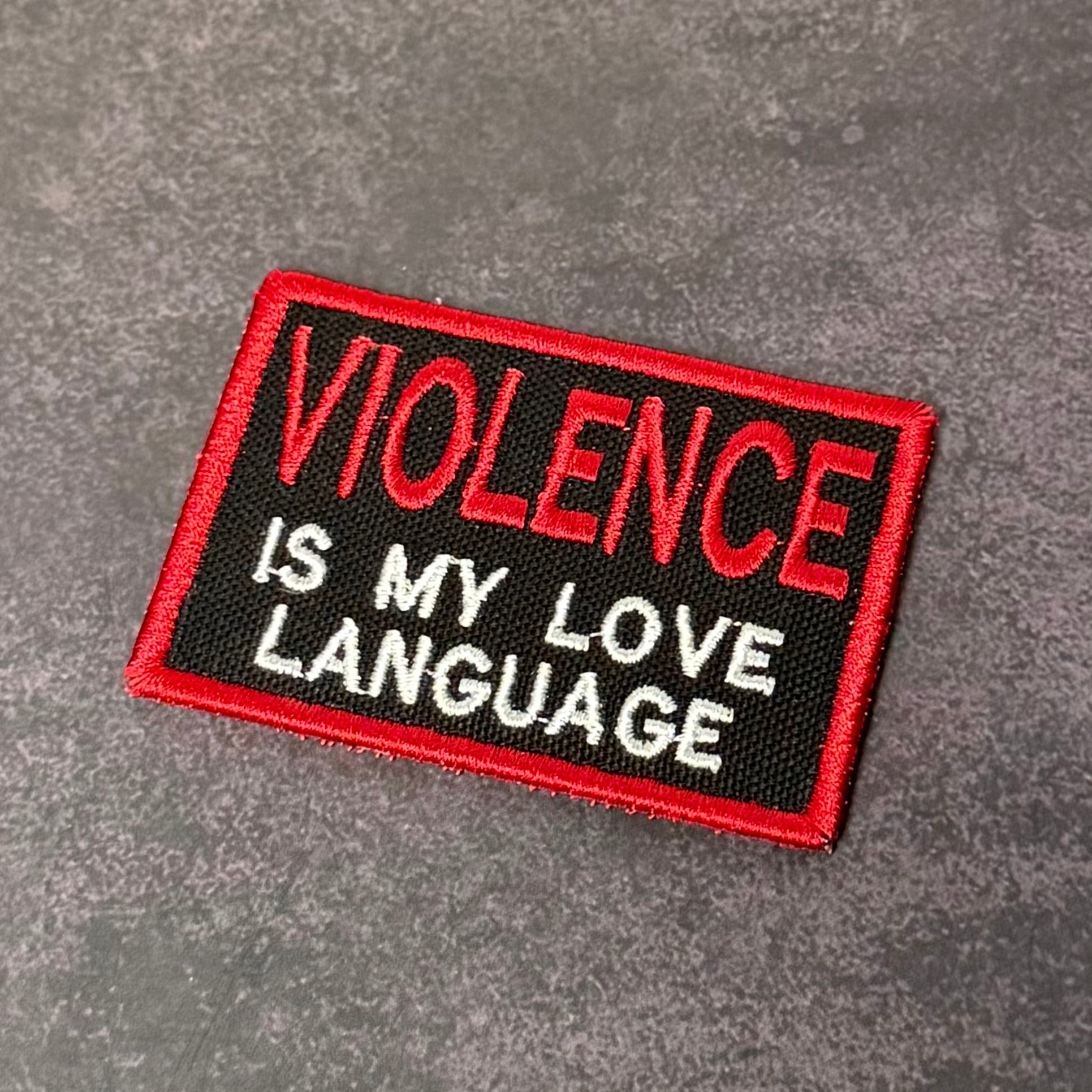 Black embroidered morale patch with red and white text reading "Violence Is My Love Language" and a red border displayed on a gray background.