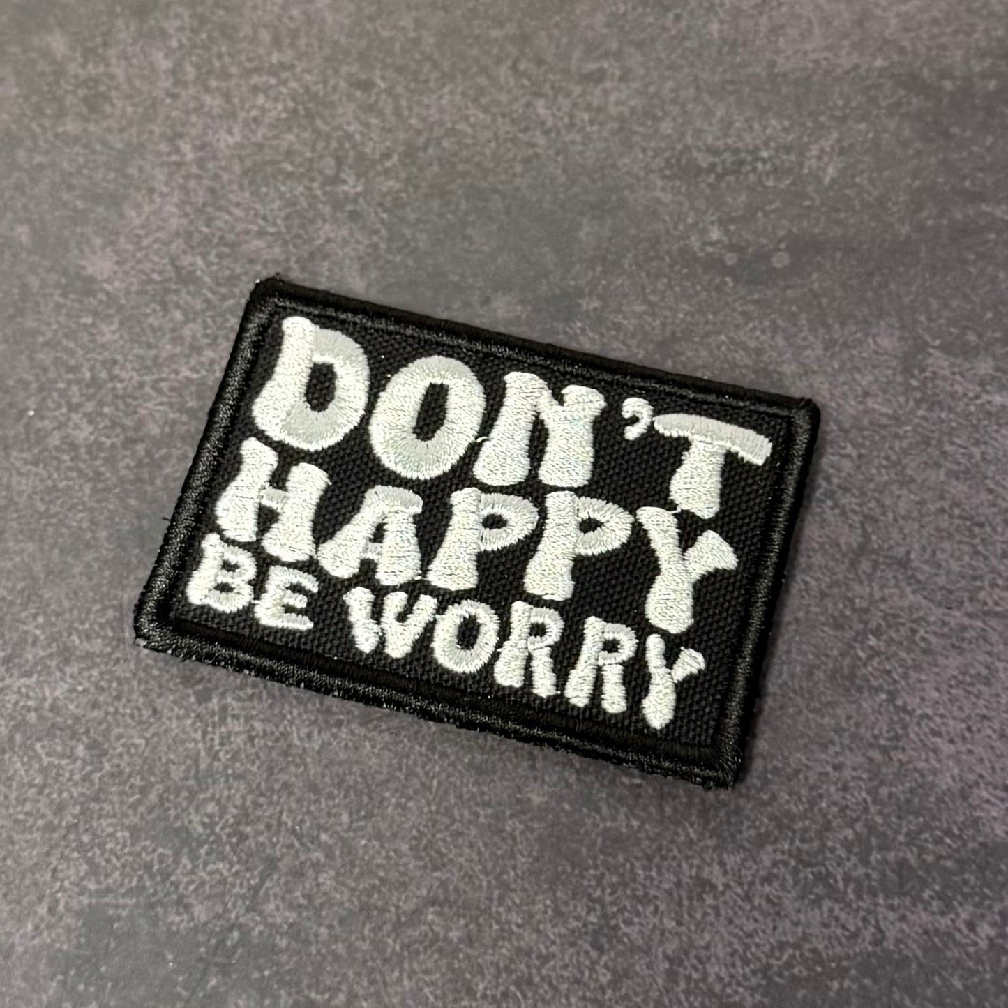 Black embroidered morale patch featuring the phrase "Don't Happy, Be Worry" in bold white lettering on a gray background.