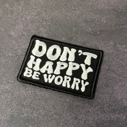 Black embroidered morale patch featuring the phrase "Don't Happy, Be Worry" in bold white lettering on a gray background.