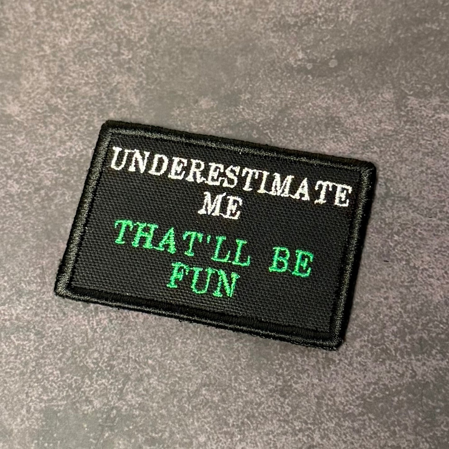Black embroidered morale patch with white and green lettering reading "Underestimate Me That'll Be Fun" displayed on a gray textured background.