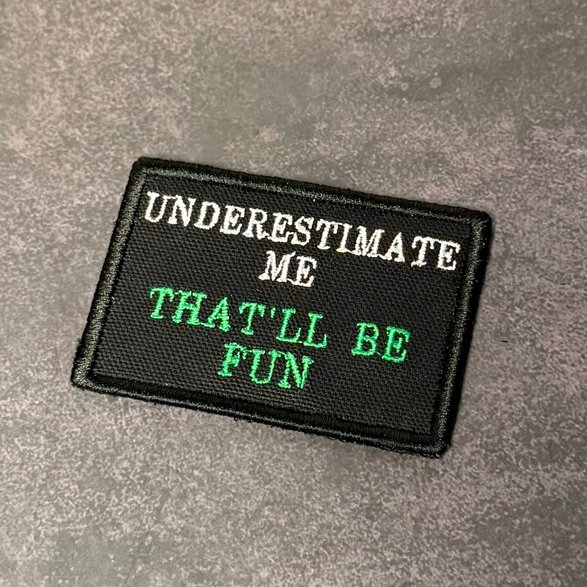 Black embroidered morale patch with white and green lettering reading "Underestimate Me That'll Be Fun" displayed on a gray textured background.