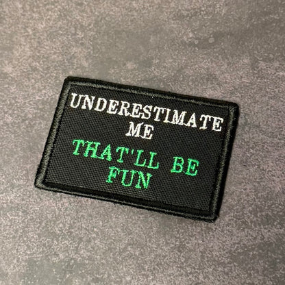 Black embroidered morale patch with white and green lettering reading "Underestimate Me That'll Be Fun" displayed on a gray textured background.