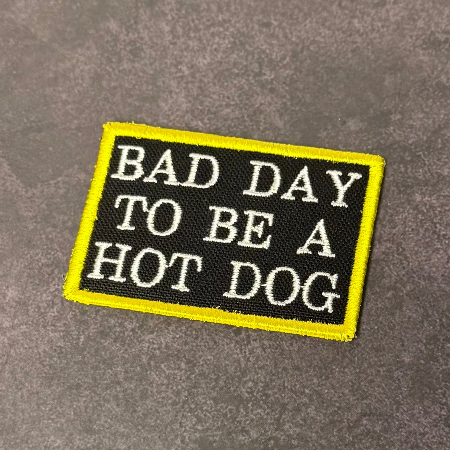 Black embroidered morale patch with white text reading "Bad Day to Be a Hot Dog" and a bright yellow border displayed on a gray background.