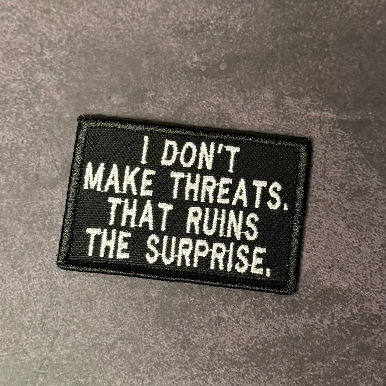 Embroidered patch with text 'I don't make threats. That ruins the surprise.' on a gray background