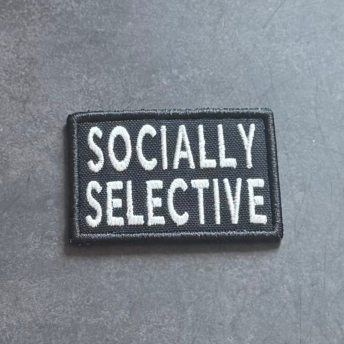 A rectangular black embroidered patch with the words 'SOCIALY SELECTIVE' in white capital letters, placed on a textured gray surface.