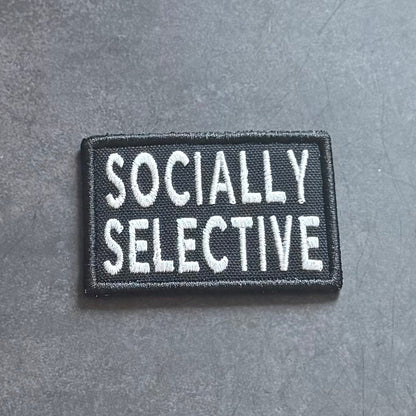 A rectangular black embroidered patch with the words 'SOCIALY SELECTIVE' in white capital letters, placed on a textured gray surface.