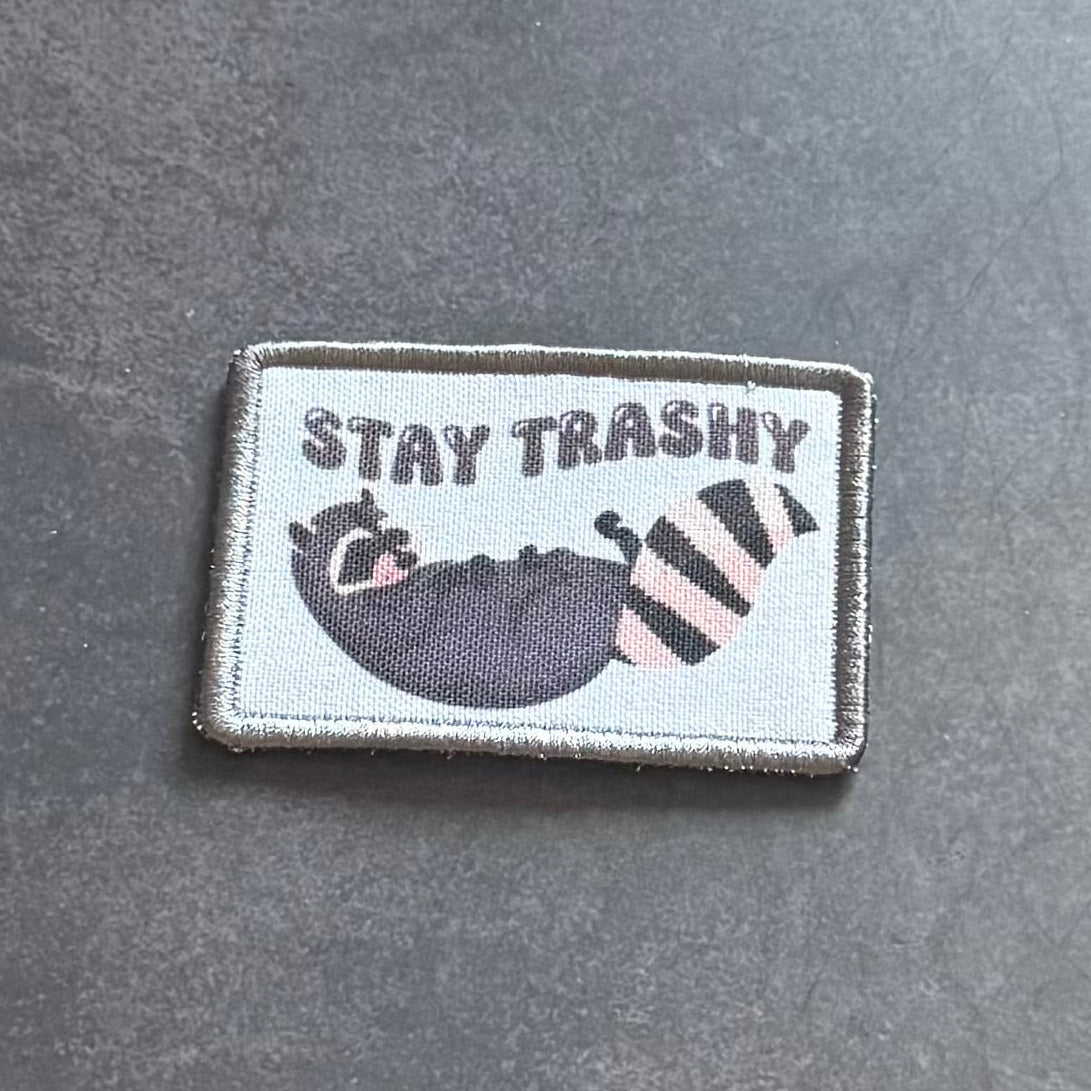 A rectangular embroidered patch with a gray border on a dark gray textured background. The patch has a light gray center with the text 'STAY TRASHY' above a cartoon raccoon in a black bowl next to a pink-and-black striped trash can.