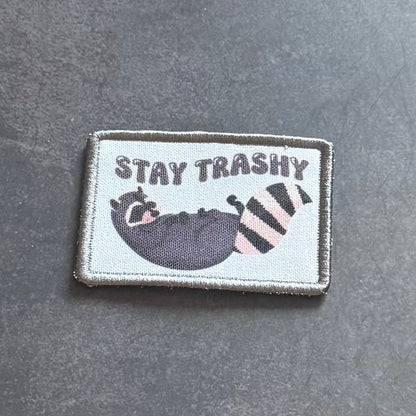 A rectangular embroidered patch with a gray border on a dark gray textured background. The patch has a light gray center with the text 'STAY TRASHY' above a cartoon raccoon in a black bowl next to a pink-and-black striped trash can.