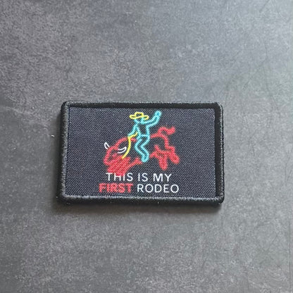 A rectangular black embroidered patch featuring a colorful graphic of a rider on a horse with the text 'THIS IS MY FIRST RODEO' in white and red, placed on a dark gray textured surface.