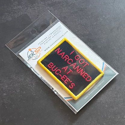 An embroidered rectangular patch with a black background, yellow border, and red text reading 'I GOT NARCANED AT BUG-EE'S', shown inside clear retail packaging on a dark gray surface.