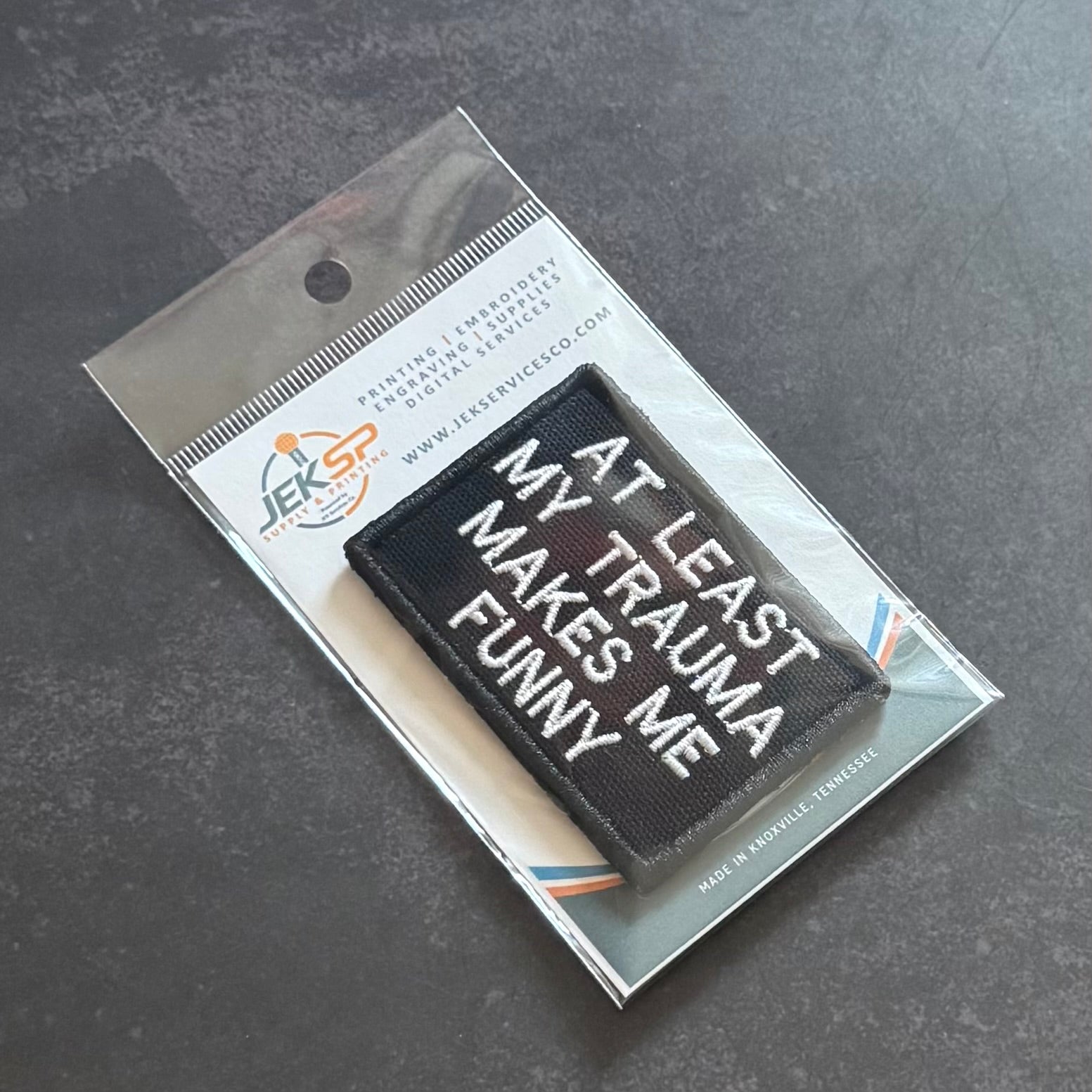 A black rectangular embroidered patch with white text reading 'AT LEAST MY TRAUMA MAKES ME FUNNY,' sealed in a clear plastic retail package on a dark gray surface.