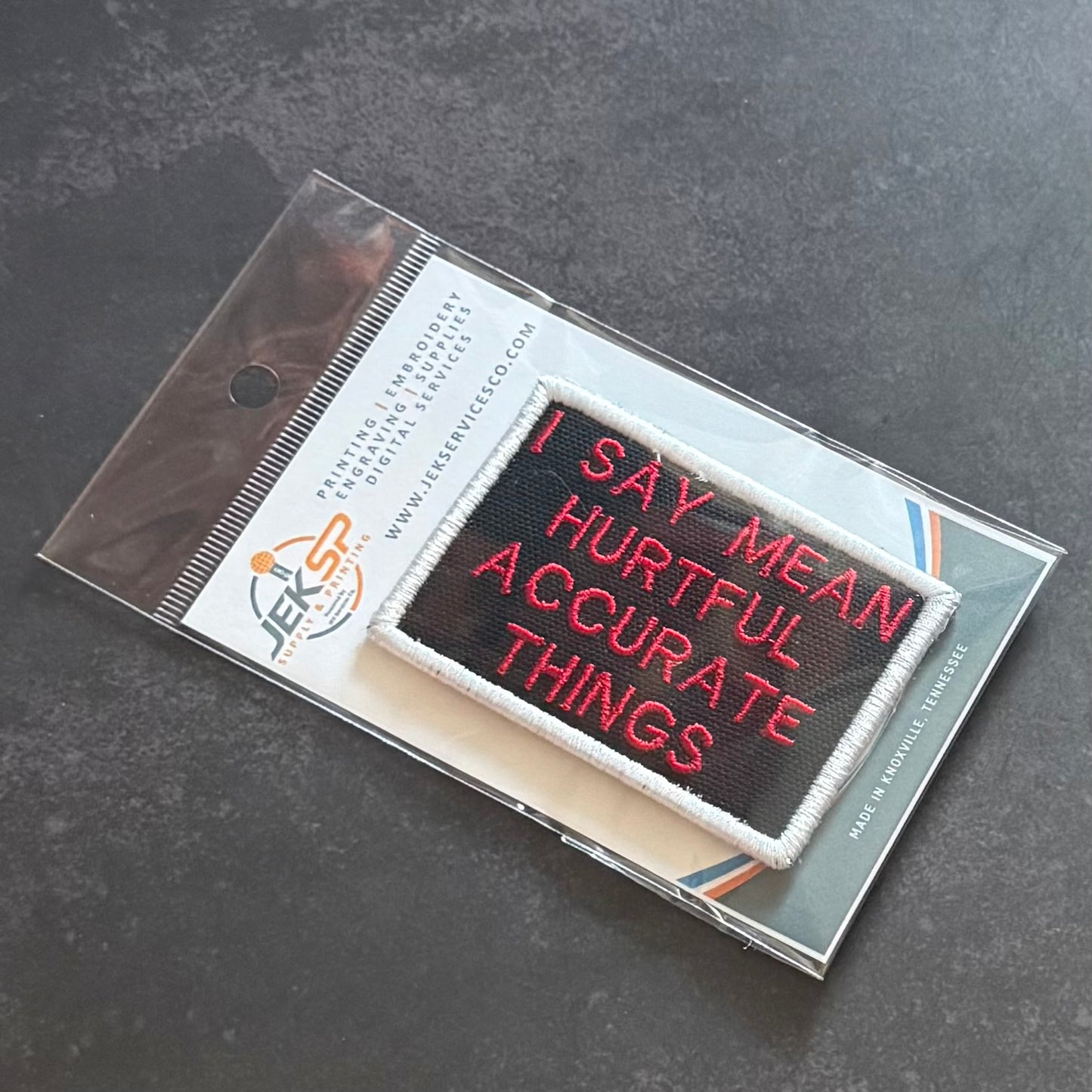 An embroidered patch with the text 'I SAY MEAN HURTFUL ACCURATE THINGS' in red on a black background with a white border, packaged in a clear plastic sleeve on a branded card.