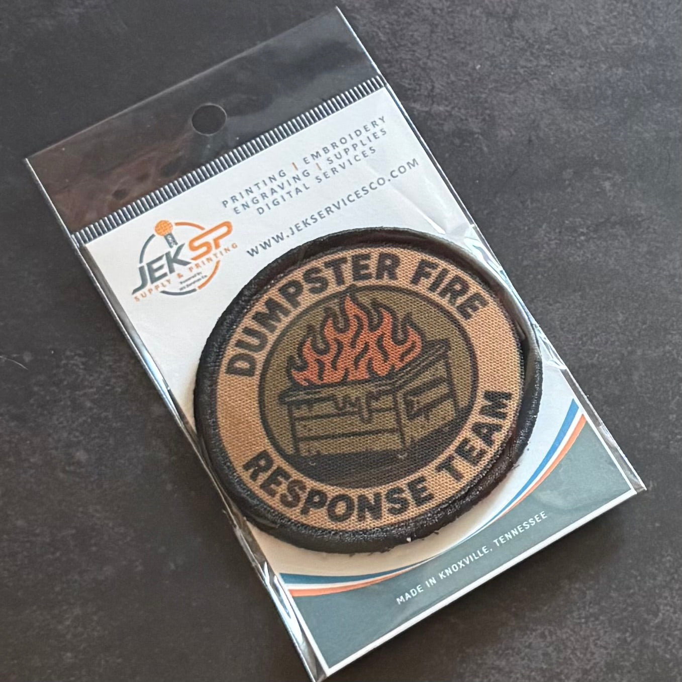 A circular embroidered patch reading 'DUMPSTER FIRE RESPONSE TEAM' with a fire graphic, shown inside clear retail packaging on a dark textured surface.